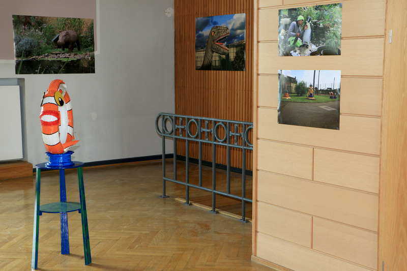 Neben der Skulptur von "Nemo" aus einem alten Autoreifen hergestellt, sind ein kleines Nilpferd vor einem Teich und der Kopf eines Dinosauriers vor einer dramatisch wirkenden Wolkenformation auf Fotografien im Hintergrund zu sehen.